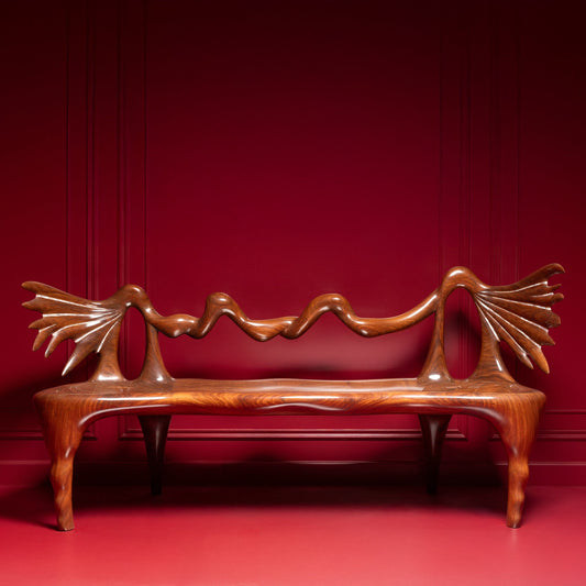 A sculptural wooden bench with a curved, organic backrest and fan-like armrests, shown against a deep red paneled wall and matching red floor.