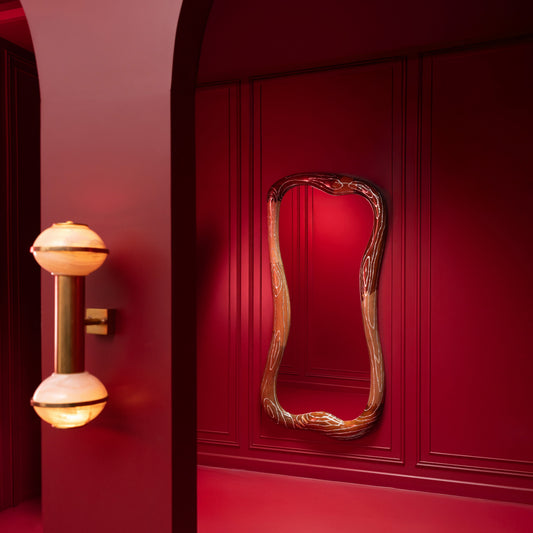 An ornate, asymmetrical oval mirror with a carved gold frame hangs on a deep red paneled wall in a matching red interior, next to a modern wall sconce.